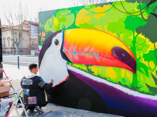 Un artiste peint un toucan coloré sur un mur, entouré de verdure.