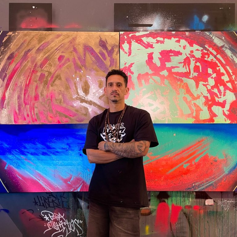 Pablito Mourer Artiste peintre Artiste de rue tenant une bombe de peinture, fond coloré et mural artistique en arrière-plan.