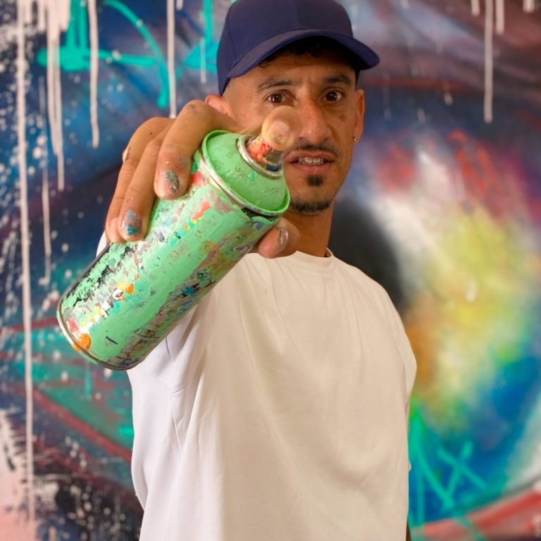 Pablito Mourer Artiste peintre Artiste de rue tenant une bombe de peinture, fond coloré et mural artistique en arrière-plan.