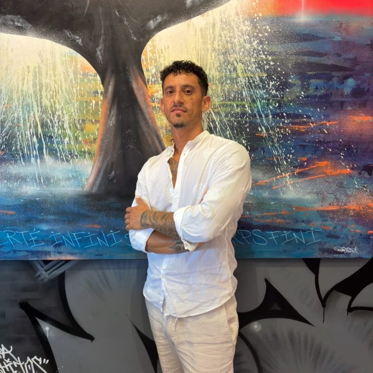 Pablito Mourer Artiste peintre Artiste de rue tenant une bombe de peinture, fond coloré et mural artistique en arrière-plan.